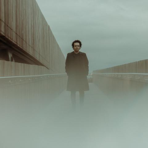 A wide shot of Maysun outdoors. There is fog, he wears a coat. 