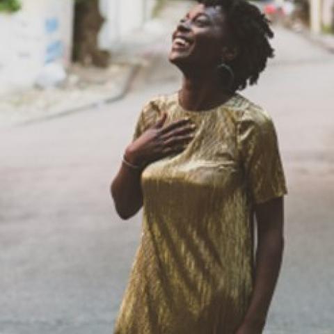 Nathalie Joachim stands smiling with her hand on her chest, in a gold dress, on a street.