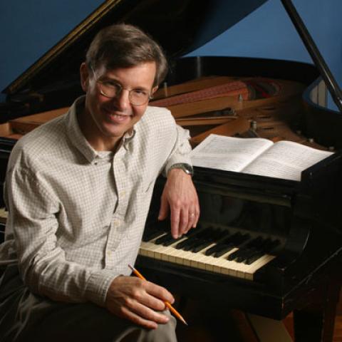 Rob Kapilow sits at a piano with a pencil in his hand and sheet music resting on top of the piano