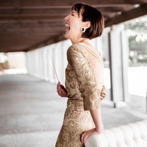 Janna stands by a white couch laughing. She wears a golden dress and holds a conductors baton. 