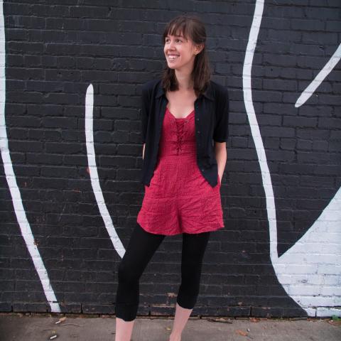 Stephanie stands with her hands in the pockets of her red and white jumper agains a black and white brick wall. 