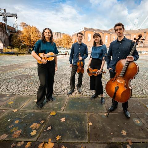 The four members of the quartet stand outdoors wearing similar outfits. they hold their instruments. 