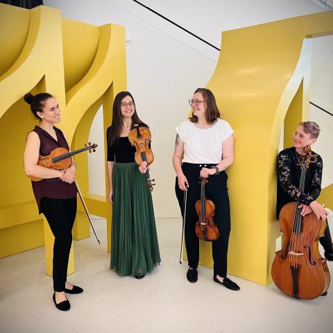 The members of the quartet stand in front of pink geometric shapes. They hold their instruments. 
