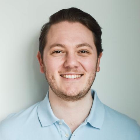 Man with short hair parted to the left smiling directly at the camera wearing a light blue polo shirt