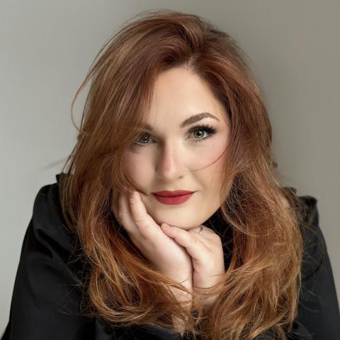 Headshot of person clasping hands under chin, red hair and lipstick 