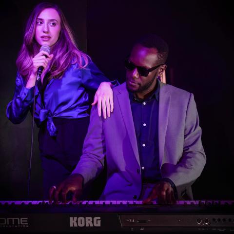 A singer holding a microphone stands beside a keyboard player performing under purple stage lighting. The keyboard player, wearing sunglasses and a light gray suit, plays a Korg keyboard while the singer leans casually on their shoulder in an intimate live music setting