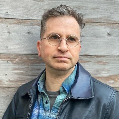 Man standing against wooden wall wearing a plaid shirt and leather jacket